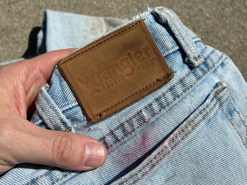 Peut inclure: Gros plan sur un jean en denim bleu clair avec une &eacute;tiquette Wrangler en cuir marron. L'&eacute;tiquette est rectangulaire avec le nom de la marque et "RUGGED WEAR" en relief. Le jean pr&eacute;sente des signes d'usure et un aspect d&eacute;lav&eacute;.