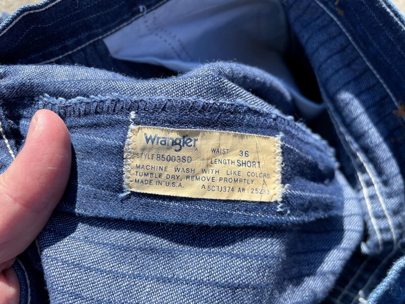 May include: Close-up of blue denim shorts with a Wrangler tag. The tag reads "WAIST 36 LENGTH SHORT" and includes washing instructions. The shorts are made in the U.S.A.