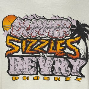 May include: White t-shirt with a graphic design that says "Summer Sizzles at DeVry Phoenix". The graphic is in a cartoon style with a sun, palm trees, and a grid pattern.