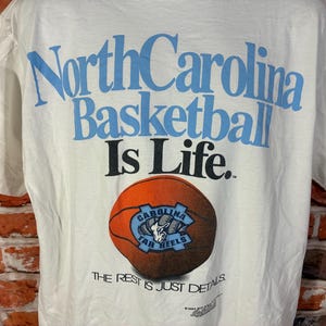 May include: White t-shirt featuring the text "North Carolina Basketball Is Life" in light blue. A basketball with the Tar Heels logo is below the text. The phrase "THE REST IS JUST DETAILS" is printed at the bottom.