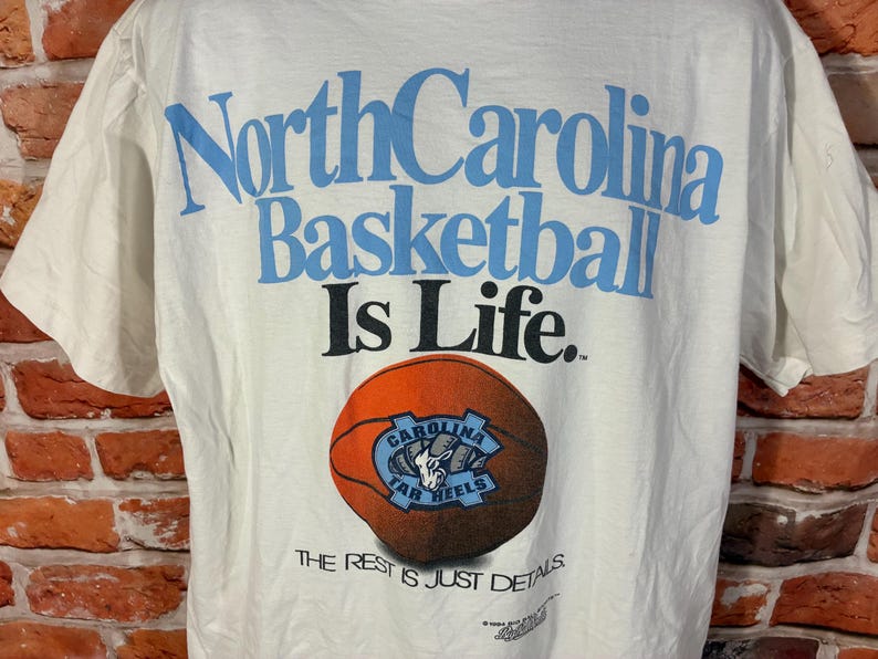 May include: White t-shirt featuring the text "North Carolina Basketball Is Life" in light blue. A basketball with the Tar Heels logo is below the text. The phrase "THE REST IS JUST DETAILS" is printed at the bottom.