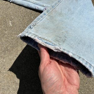 Peut inclure: Un jean en denim bleu clair avec un ourlet effiloch&eacute; et vieilli. Le jean est tenu par une main, montrant le tissu us&eacute; et d&eacute;lav&eacute;. Le jean est probablement vintage ou a un aspect us&eacute;.