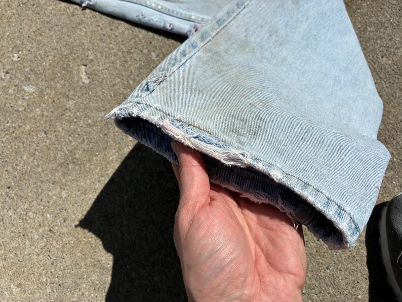 Peut inclure: Un jean en denim bleu clair avec un ourlet effiloch&eacute; et vieilli. Le jean est tenu par une main, montrant le tissu us&eacute; et d&eacute;lav&eacute;. Le jean est probablement vintage ou a un aspect us&eacute;.