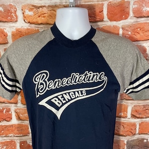 May include: A navy blue and gray baseball t-shirt with the text "Benedictine Bengals" in white.