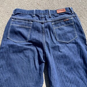 May include: Close-up of the back of blue denim jeans with vertical white pinstripes. The jeans have two back pockets with white stitching, a brown leather patch, and a small tag. The overall style is vintage.