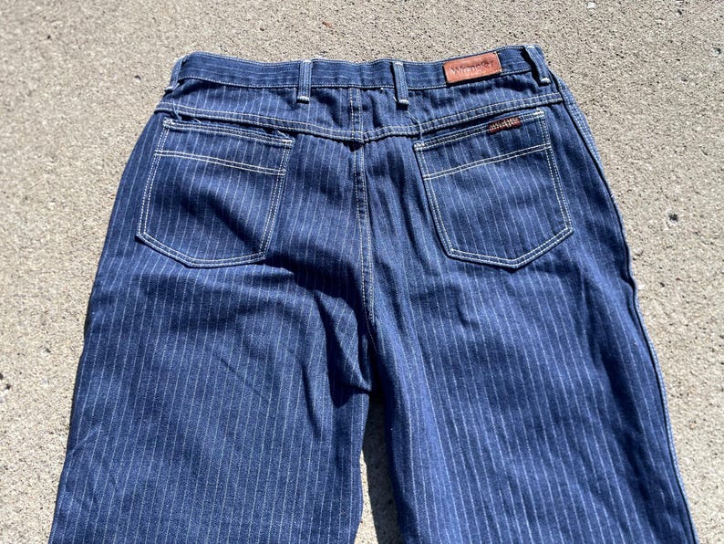 May include: Close-up of the back of blue denim jeans with vertical white pinstripes. The jeans have two back pockets with white stitching, a brown leather patch, and a small tag. The overall style is vintage.