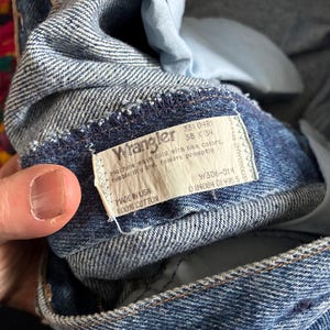 May include: Close-up of blue denim Wrangler jeans. The label reads "Wrangler" with details including size 38 x 34, and "Made in USA 100% Cotton." The jeans are folded, showing the inside lining and stitching.