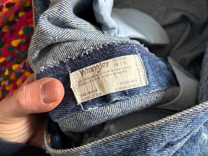 May include: Close-up of blue denim Wrangler jeans. The label reads "Wrangler" with details including size 38 x 34, and "Made in USA 100% Cotton." The jeans are folded, showing the inside lining and stitching.