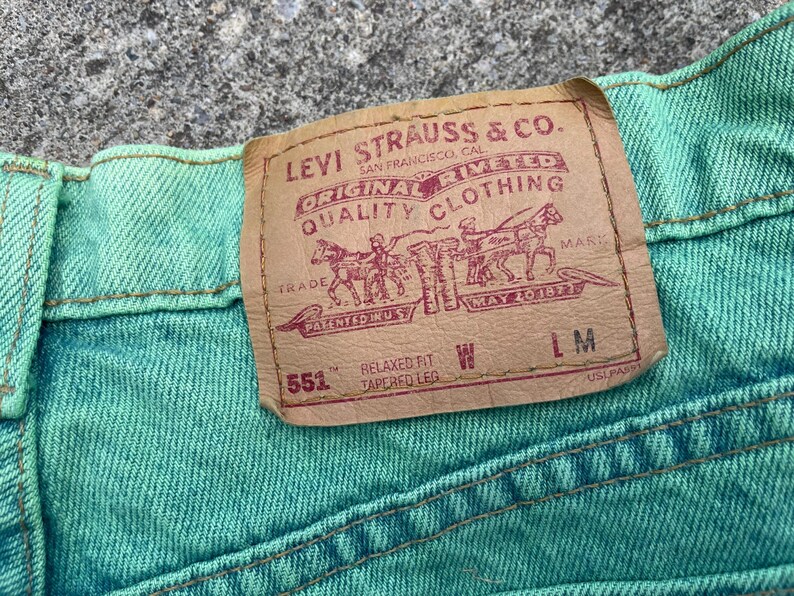 May include: Close-up of a section of turquoise denim jeans. A rectangular brown leather patch with the Levi Strauss & Co. logo is sewn onto the jeans. The patch includes text and a graphic of people and horses.