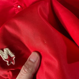 May include: Bright red satin jacket with a small embroidered patch featuring a white letter "M" and a red star. The jacket has a smooth, shiny texture and a classic design.