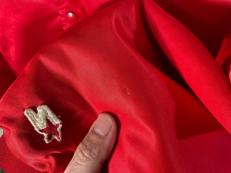 May include: Bright red satin jacket with a small embroidered patch featuring a white letter "M" and a red star. The jacket has a smooth, shiny texture and a classic design.