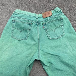 May include: Back view of green denim jeans with classic five-pocket styling. The jeans feature a brown leather patch and a red Levi's tag on the back pocket. The jeans are made of a durable denim fabric.