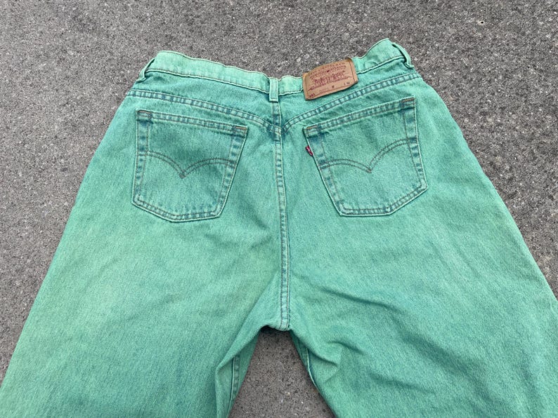 May include: Back view of green denim jeans with classic five-pocket styling. The jeans feature a brown leather patch and a red Levi's tag on the back pocket. The jeans are made of a durable denim fabric.