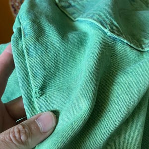 May include: Close-up of a distressed, light green denim fabric. The fabric has a textured appearance with visible diagonal weave lines. There is a small tear in the fabric, adding to the worn aesthetic. The denim appears to be part of a garment.