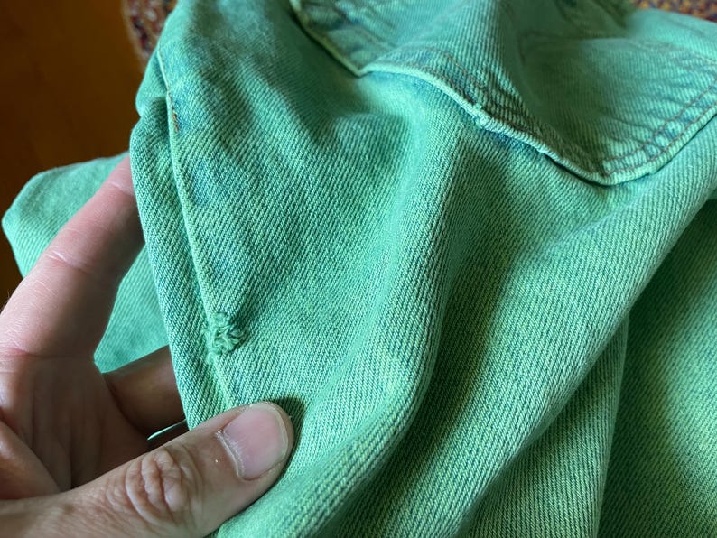 May include: Close-up of a distressed, light green denim fabric. The fabric has a textured appearance with visible diagonal weave lines. There is a small tear in the fabric, adding to the worn aesthetic. The denim appears to be part of a garment.