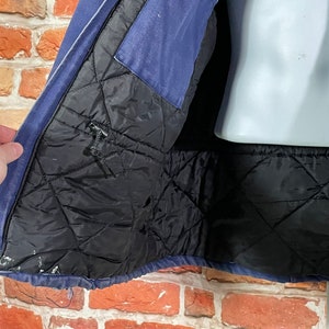 May include: A blue denim jacket with a black quilted lining. The jacket is unzipped and the lining is visible. The jacket has a zipper closure and a pocket on the left side.
