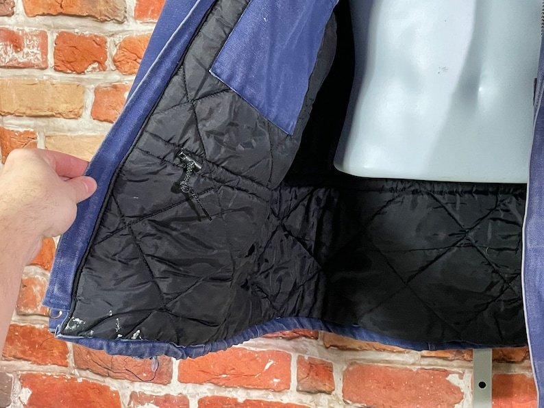 May include: A blue denim jacket with a black quilted lining. The jacket is unzipped and the lining is visible. The jacket has a zipper closure and a pocket on the left side.