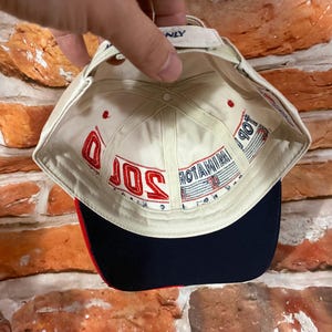 Op de afbeelding: Een gebroken witte baseballpet met een donkerblauwe klep en rode accenten. De binnenkant is voorzien van rood geborduurde tekst 2000 en blauwe tekst TERMINATOR. De pet heeft een verstelbare band aan de achterkant met een klein blauw label.