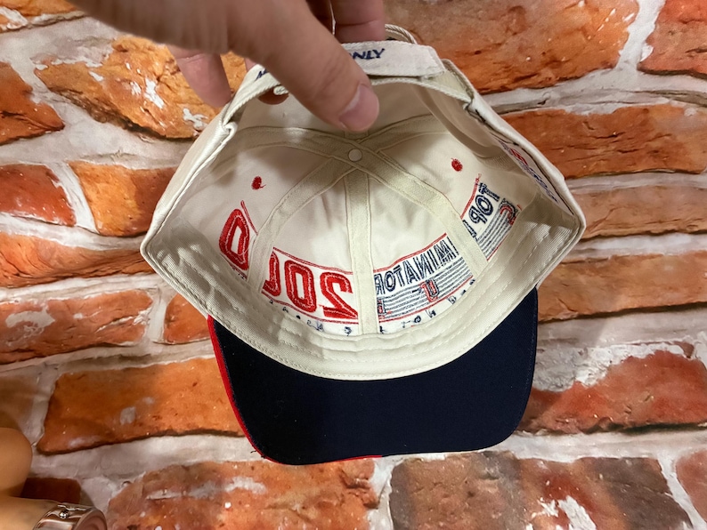 Op de afbeelding: Een gebroken witte baseballpet met een donkerblauwe klep en rode accenten. De binnenkant is voorzien van rood geborduurde tekst 2000 en blauwe tekst TERMINATOR. De pet heeft een verstelbare band aan de achterkant met een klein blauw label.
