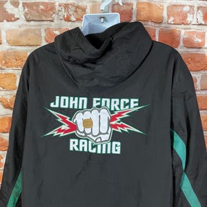 May include: Black hooded jacket with green accents and the text "JOHN FORCE RACING" in white and green. The design includes a fist graphic with a ring and lightning bolts.