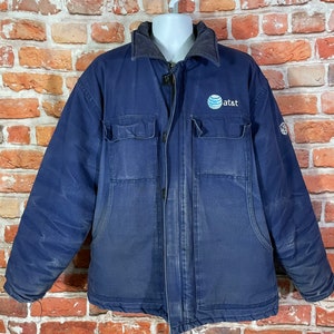 May include: A dark blue work jacket with a zipper closure and a large "at&t" logo on the chest. The jacket has two large pockets on the front and a smaller pocket on the left sleeve.