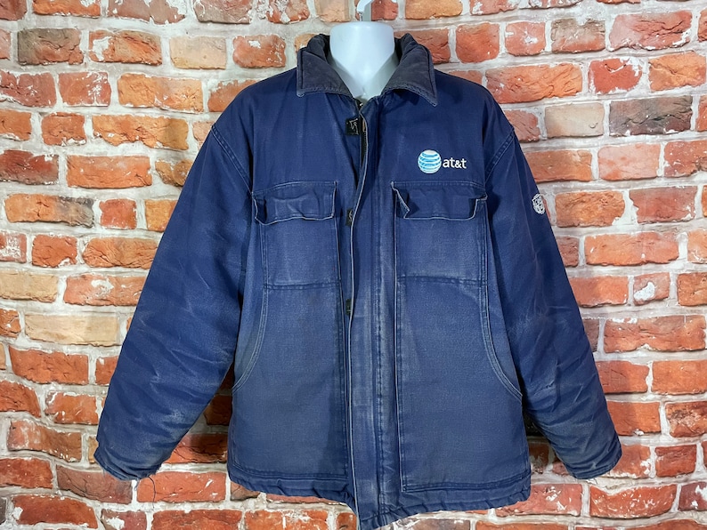 May include: A dark blue work jacket with a zipper closure and a large "at&t" logo on the chest. The jacket has two large pockets on the front and a smaller pocket on the left sleeve.