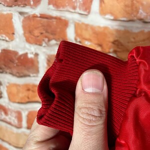 May include: Close-up of a red ribbed knit cuff and a red satin fabric. The cuff is held by a hand, revealing the texture and color of the material. The background is a brick wall.
