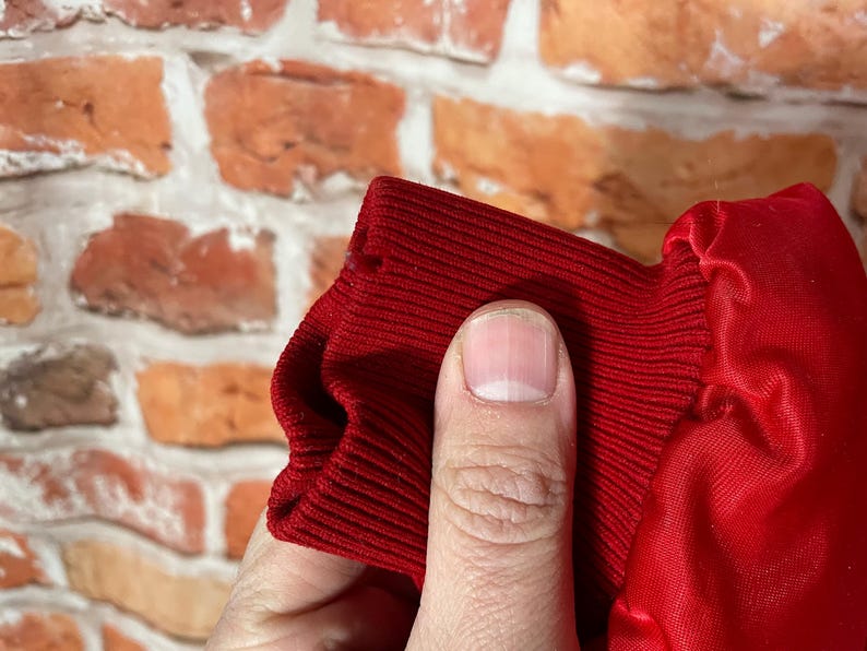 May include: Close-up of a red ribbed knit cuff and a red satin fabric. The cuff is held by a hand, revealing the texture and color of the material. The background is a brick wall.