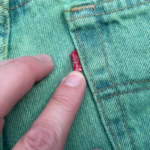 May include: Close-up of green denim fabric with visible stitching and a red Levi's tag. The fabric is a light green color, and the stitching is a contrasting brown. The tag is red with white text.