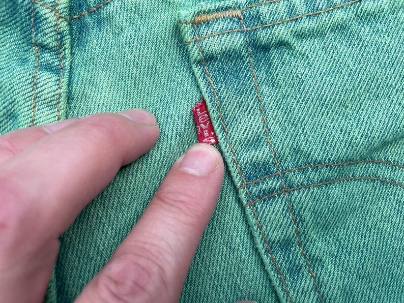 May include: Close-up of green denim fabric with visible stitching and a red Levi's tag. The fabric is a light green color, and the stitching is a contrasting brown. The tag is red with white text.