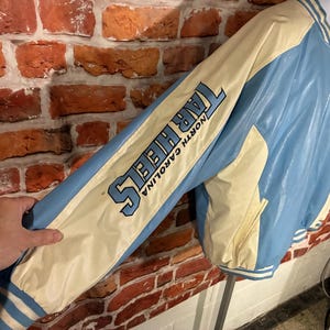 May include: A light blue and cream-colored Tar Heels jacket with the words "TAR HEELS NORTH CAROLINA" printed on the sleeve. The jacket has a zipper closure and a white and blue striped trim.