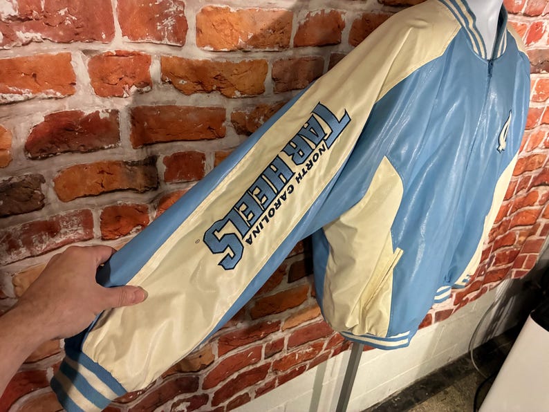 May include: A light blue and cream-colored Tar Heels jacket with the words "TAR HEELS NORTH CAROLINA" printed on the sleeve. The jacket has a zipper closure and a white and blue striped trim.