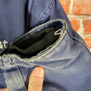 May include: Close-up of the navy blue sleeve of a jacket with a black ribbed knit cuff. The cuff is frayed and worn. The jacket has a white embroidered "at&t" logo on the front.