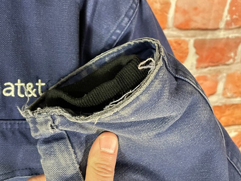 May include: Close-up of the navy blue sleeve of a jacket with a black ribbed knit cuff. The cuff is frayed and worn. The jacket has a white embroidered "at&t" logo on the front.
