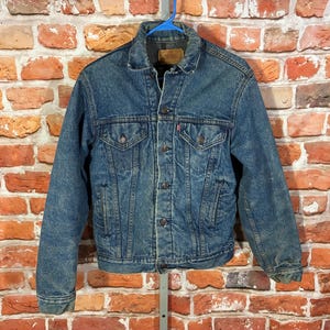 May include: A medium-wash blue denim jacket featuring a pointed collar, long sleeves with buttoned cuffs, and a front button closure. The jacket includes two chest pockets with buttoned flaps, two side pockets, and a small red brand tab on the left pocket.