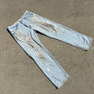 Peut inclure: Jean en denim bleu clair avec des d&eacute;tails us&eacute;s, notamment des d&eacute;chirures et des taches. Le jean est pos&eacute; &agrave; plat, mettant en valeur les poches avant et arri&egrave;re, et l'aspect g&eacute;n&eacute;ral us&eacute;.