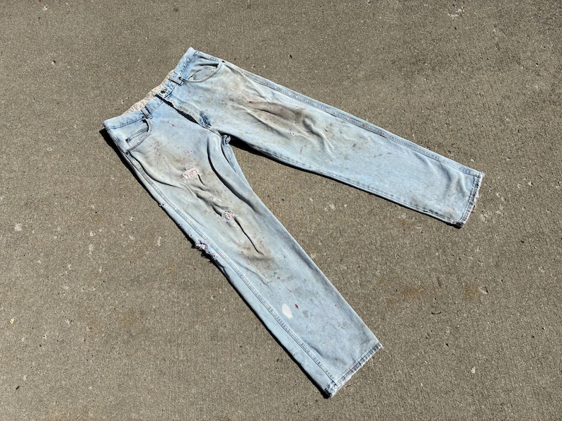 Peut inclure: Jean en denim bleu clair avec des d&eacute;tails us&eacute;s, notamment des d&eacute;chirures et des taches. Le jean est pos&eacute; &agrave; plat, mettant en valeur les poches avant et arri&egrave;re, et l'aspect g&eacute;n&eacute;ral us&eacute;.