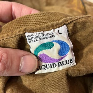 Puede incluir: Primer plano de una etiqueta de camiseta marr&oacute;n con el logotipo de Liquid Blue. La etiqueta es blanca con un dise&ntilde;o turquesa, morado y amarillo. El texto de la etiqueta dice "100% Algod&oacute;n Ensamblado en M&eacute;xico de Componentes de EE.UU." y "L".