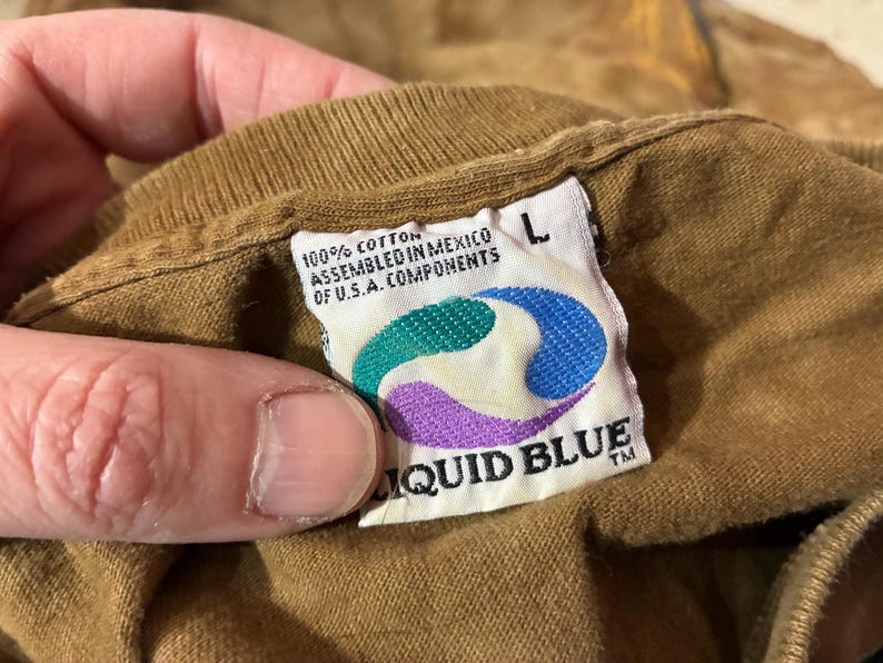 Puede incluir: Primer plano de una etiqueta de camiseta marr&oacute;n con el logotipo de Liquid Blue. La etiqueta es blanca con un dise&ntilde;o turquesa, morado y amarillo. El texto de la etiqueta dice "100% Algod&oacute;n Ensamblado en M&eacute;xico de Componentes de EE.UU." y "L".