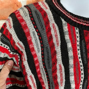Peut inclure: Un pull ray&eacute; avec des rayures noires, rouges et grises. Le pull est en tricot textur&eacute; et a un col rond.
