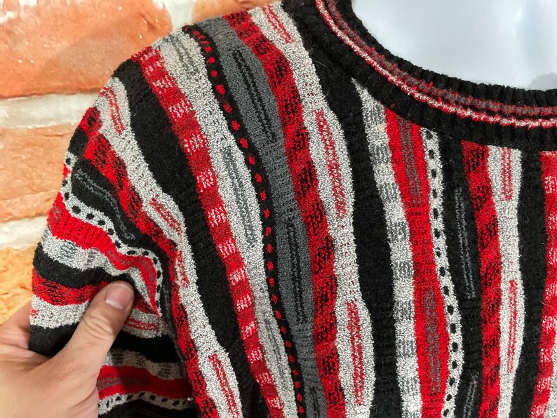 Peut inclure: Un pull ray&eacute; avec des rayures noires, rouges et grises. Le pull est en tricot textur&eacute; et a un col rond.