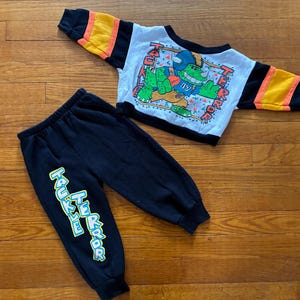 May include: A two-piece children's outfit featuring a white and black long-sleeved top and black sweatpants. The top has a graphic of a green dinosaur with the text "TAKE TERROR". The sweatpants have the same text in a vertical design.