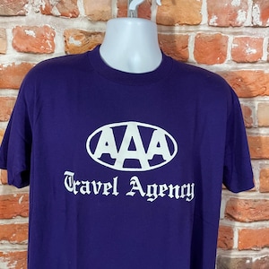 May include: Purple t-shirt with a white AAA Travel Agency logo. The logo is a white oval with the letters AAA in white inside the oval.