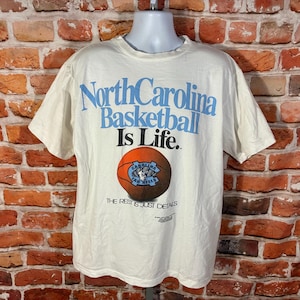 May include: Cream-colored t-shirt with the text "North Carolina Basketball Is Life." A basketball graphic is below the text. The shirt has short sleeves and a crew neck.