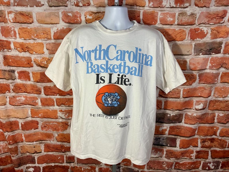May include: Cream-colored t-shirt with the text "North Carolina Basketball Is Life." A basketball graphic is below the text. The shirt has short sleeves and a crew neck.