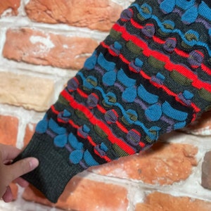 May include: A close-up of a colorful knitted sweater sleeve with a textured pattern of blue, red, green, and black. The sweater has a striped design with a repeating pattern of small circles.