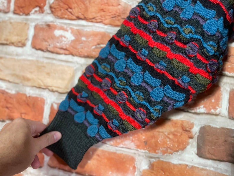 May include: A close-up of a colorful knitted sweater sleeve with a textured pattern of blue, red, green, and black. The sweater has a striped design with a repeating pattern of small circles.