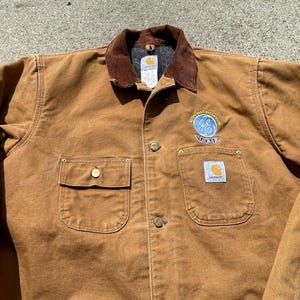 May include: A brown Carhartt work jacket with a corduroy collar and a General Electric logo patch. The jacket has a large front pocket and two smaller pockets on the chest.
