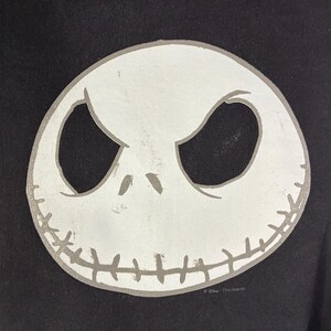 May include: Black sweatshirt featuring a large, white, stylized image of Jack Skellington's face. The design includes large oval eyes, a stitched mouth, and a gray outline. The sweatshirt is a dark color, and the design is centered.