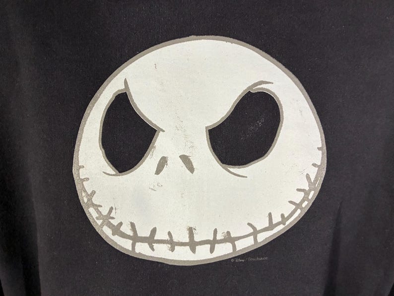 May include: Black sweatshirt featuring a large, white, stylized image of Jack Skellington's face. The design includes large oval eyes, a stitched mouth, and a gray outline. The sweatshirt is a dark color, and the design is centered.